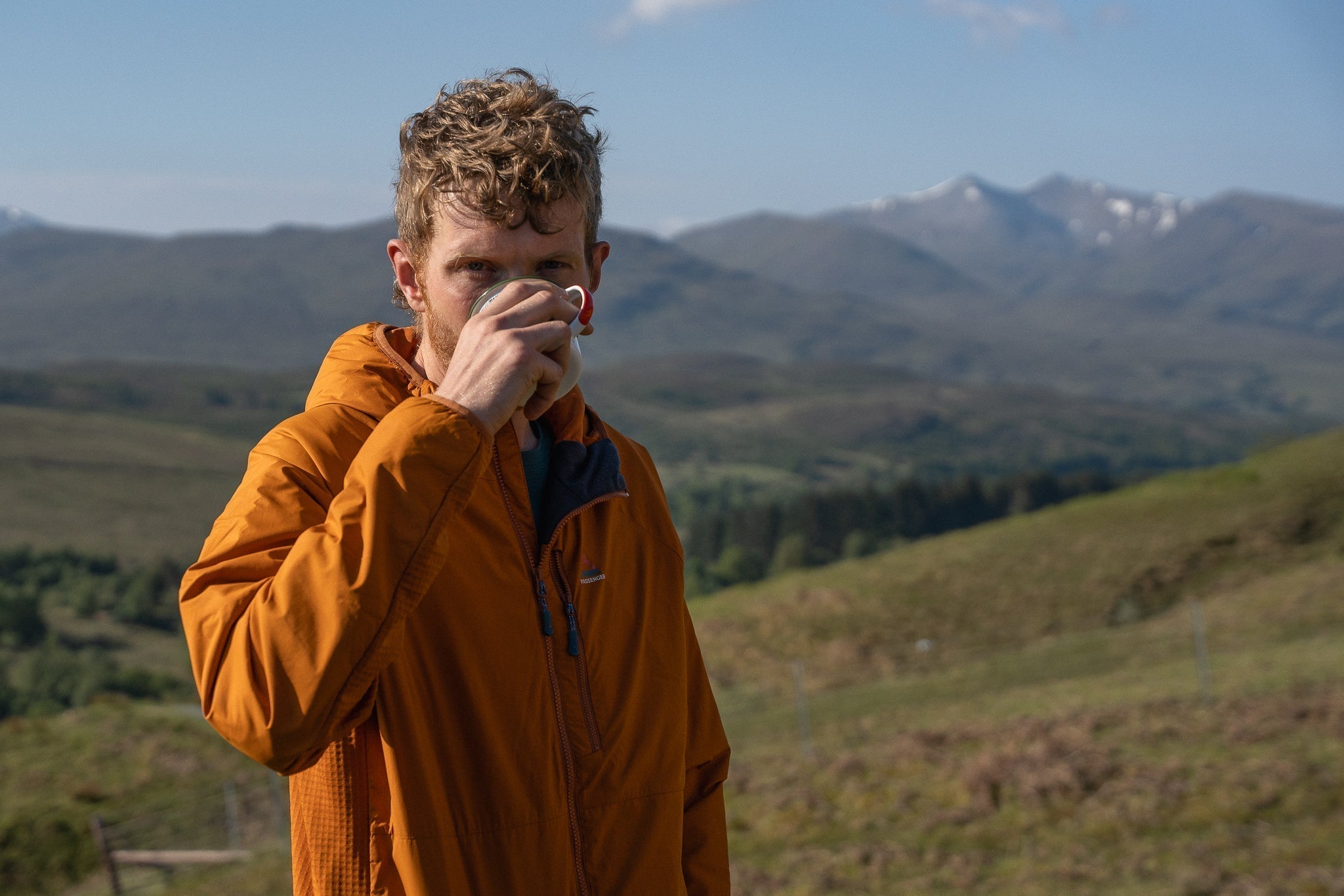 A man sipping from a mug in the Scottish Highlands wearing an orange jacket