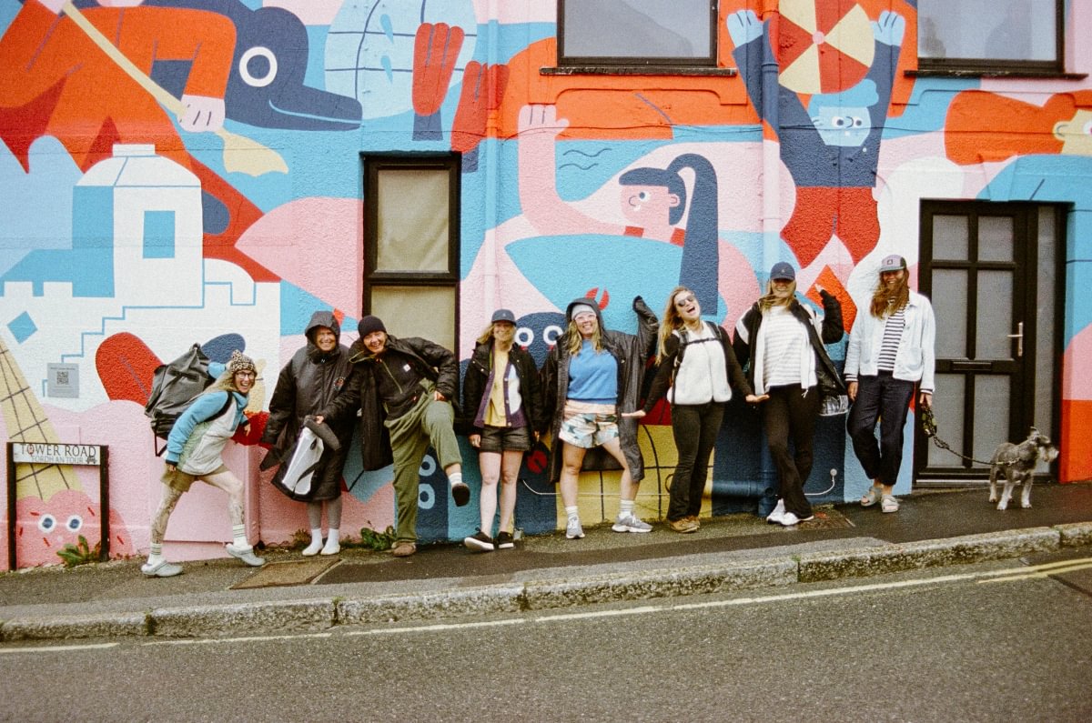 A group of eight people and a dog standing on a street curb in front of a brightly painted mural wall. The mural features abstract cartoon-like figures and shapes in orange, blue, pink, and white.