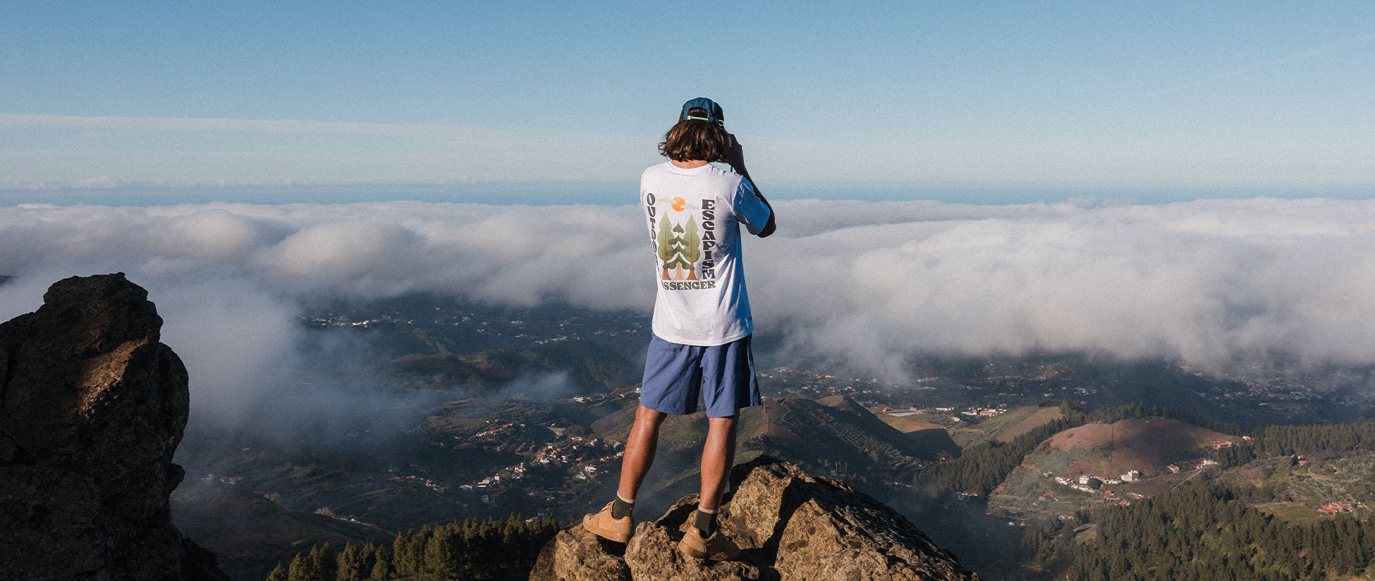 A man stands on a rock on top of a mountain, faces away from the camera and takes a photo of the view below